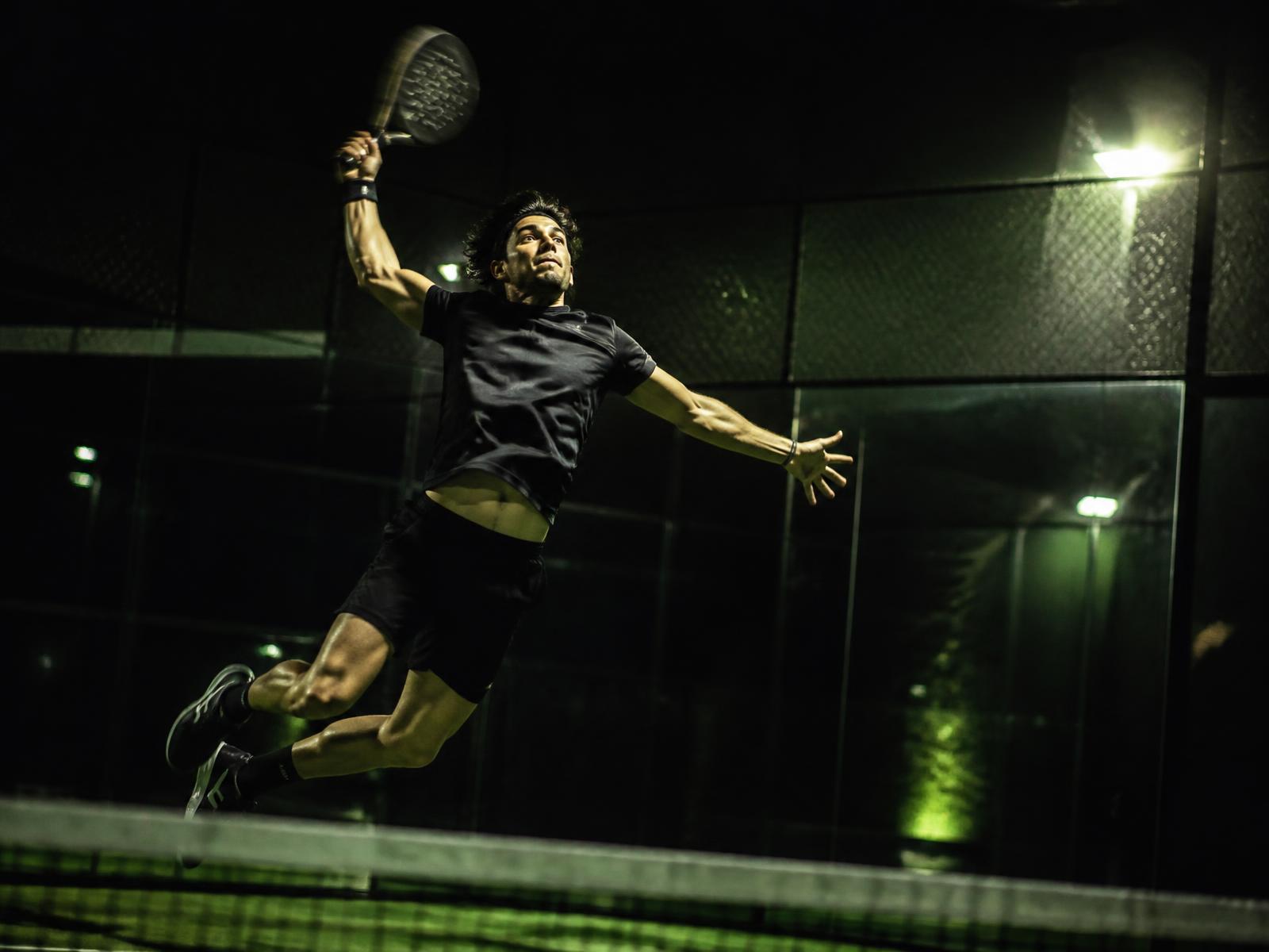 Padel player jumping to hit a smash on a floodlit glass court